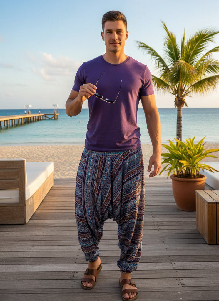 Man standing on a wooden deck by the beach holding sunglasses, wearing a purple shirt and patterned pants.